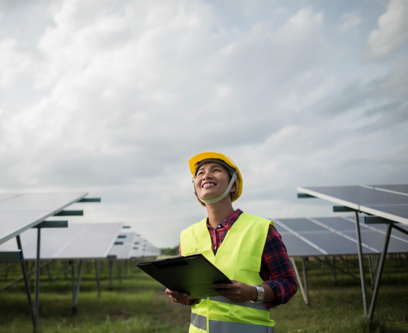 Engineer in a hard hat and safety vest holding a clipboard between rows of solar panels